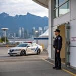 A uniformed security guard stands at a building entrance while a marked patrol car is parked nearby with the Vancouver skyline and mountains in the background.